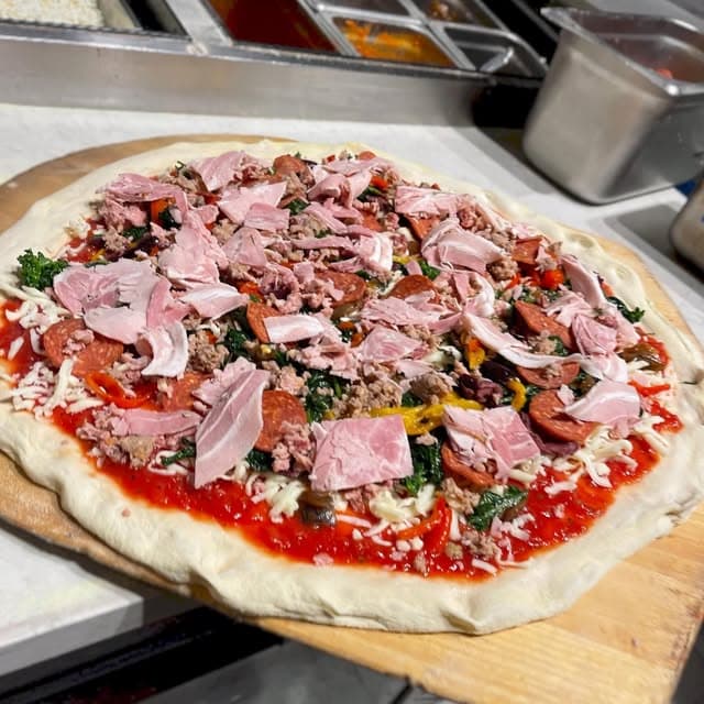 A pie being built on the peel with sausage, pepperoni, prosciutto and greens